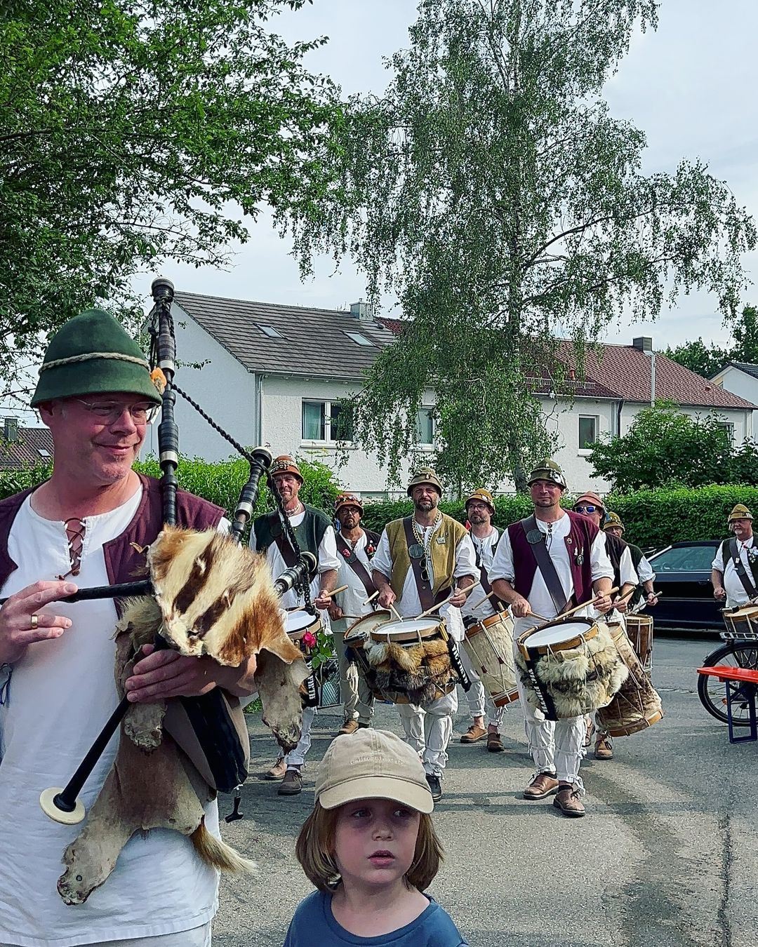 Eine Gruppe von Männern mit Trommeln und Dudelsack in traditioneller Kleidung laufen musizierend durch eine Wohngegend